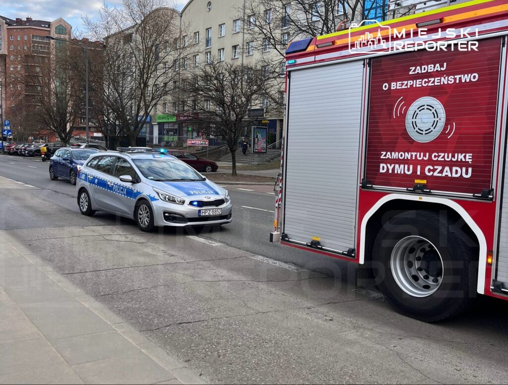 Policyjna radiowóz z włączonymi sygnałami świetlnymi porusza się po ulicy obok wozu strażackiego z napisem "Zamontuj czujkę dymu i czadu".