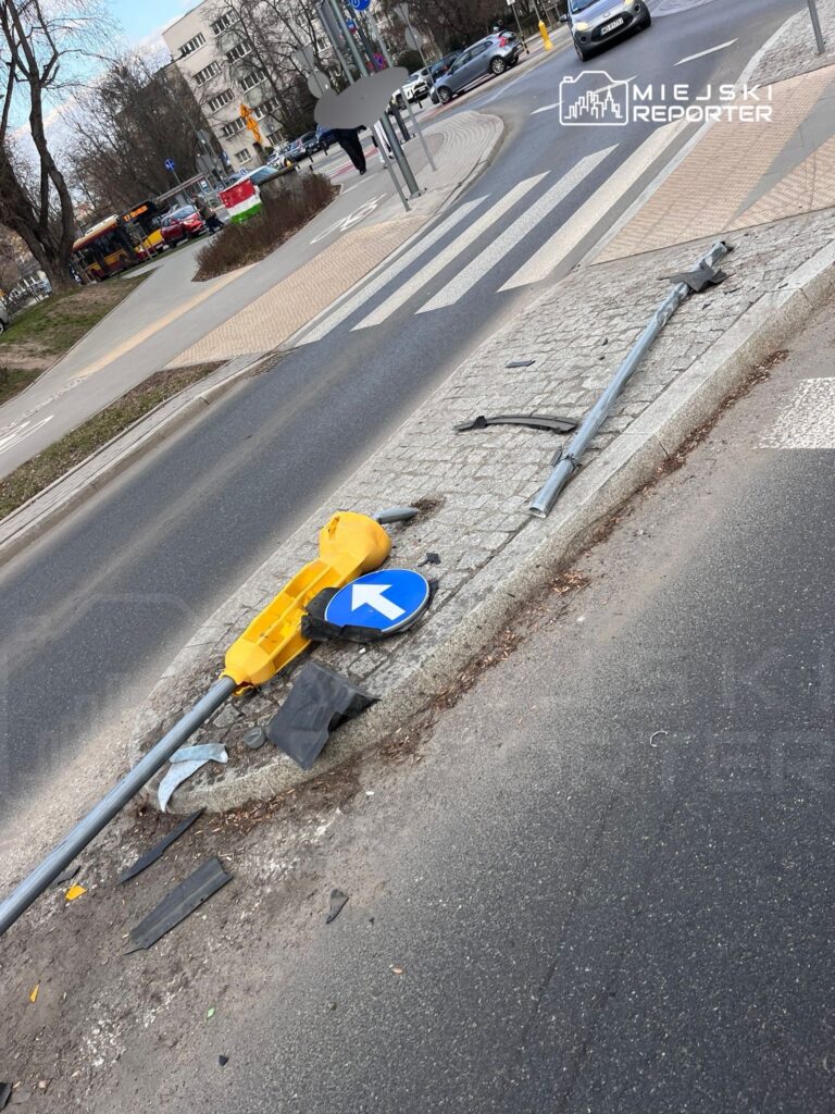 Złamany znak drogowy z kierunkowskazem leży na chodniku obok uszkodzonego słupa, w ruchliwej okolicy.