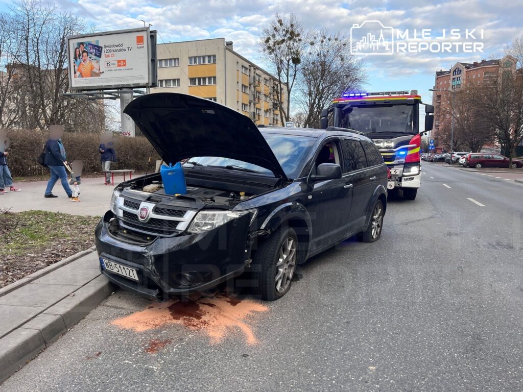 Czarny samochód osobowy z otwartą maską, w tle straż pożarna oraz przechodnie na chodniku obok.