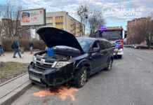 Czarny samochód osobowy z otwartą maską, w tle straż pożarna oraz przechodnie na chodniku obok.