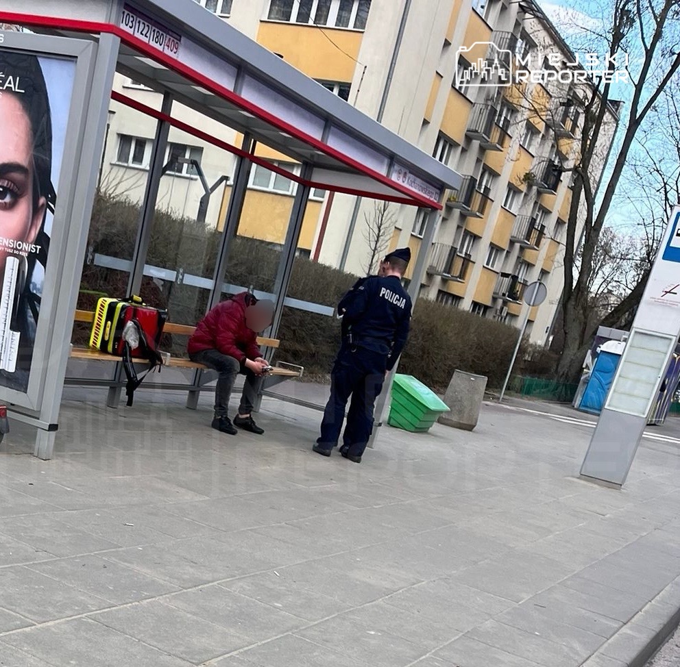Mężczyzna w czerwonej kurtce siedzi na ławce przystanku, rozmawiając z policjantem w uniformie.
