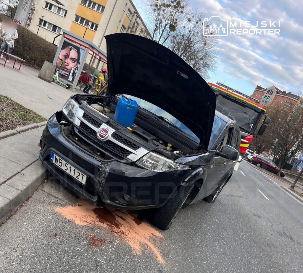 Czarny samochód osobowy z otwartą maską stoi na poboczu, z plamą cieczy na jezdni; w tle widać woz strażacki.