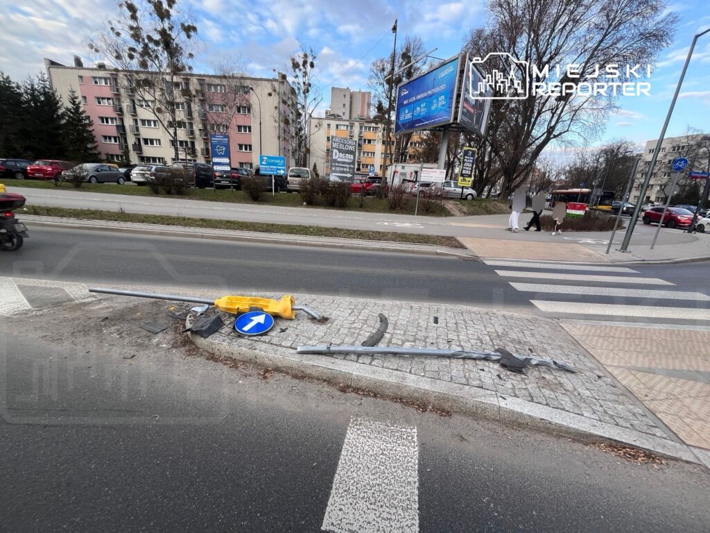 Złamany znak drogowy oraz uszkodzona sygnalizacja świetlna leżą na chodniku przy ruchliwej ulicy. W tle widoczne bloki mieszkalne i samochody.