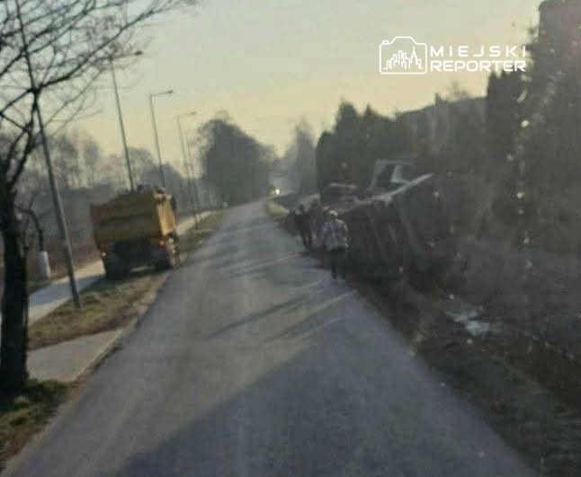Na drodze leży przewrócony pojazd, obok grupka ludzi oraz żółty samochód ciężarowy.