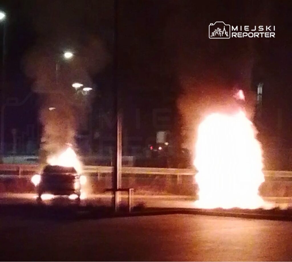 Dwa płonące samochody na parkingu, emitujące dym i ogień w nocy, otoczone słabym oświetleniem ulicznym.