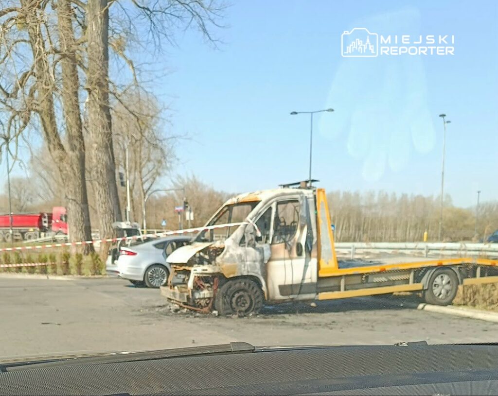 Spalony samochód dostawczy stoi na parkingu, otoczony taśmą ostrzegawczą, w tle widać inne pojazdy.