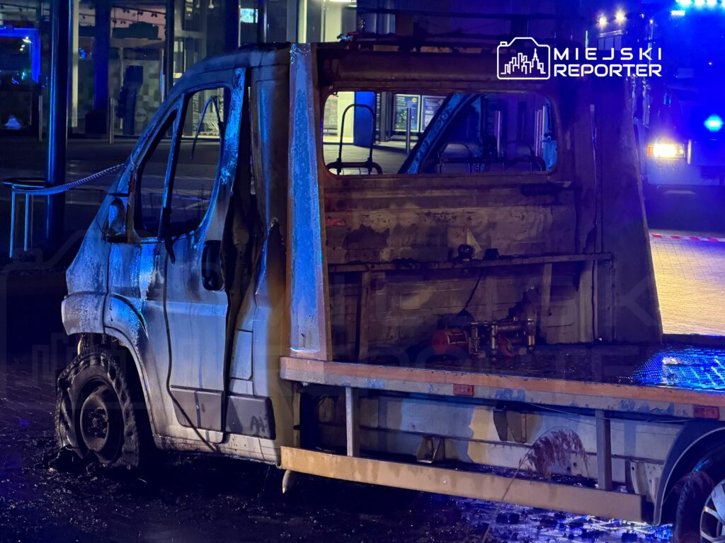 Spalony dostawczy samochód z uszkodzoną oponą stoi na mokrej nawierzchni, w tle migają niebieskie światła.
