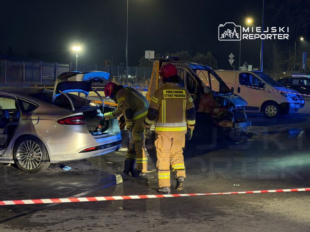 Strażacy w mundurach przeszukują bagażnik uszkodzonego samochodu na oświetlonym parkingu nocą.