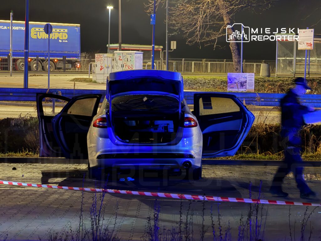 Otwarte drzwi samochodu osobowego na miejscu zdarzenia, obok policyjny funkcjonariusz przechodzący obok taśmy zabezpieczającej.