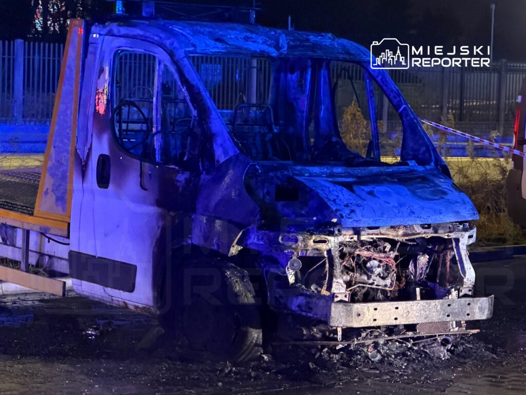 Spalona furgonetka na drodze, otoczona przez niebieskie światła policyjne i taśmy zabezpieczające.