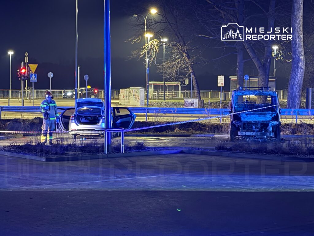 Strażak w odblaskowym mundurze bada uszkodzony samochód osobowy na oświetlonym parkingu nocą. W tle stoi spalone auto.