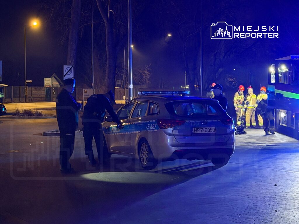 Dwaj policjanci sprawdzają samochód policyjny w nocnym otoczeniu, w tle strażacy przygotowują się do akcji.