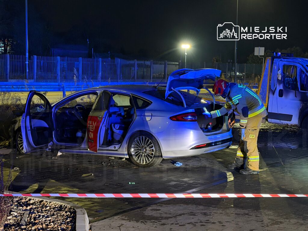 Strażak w mundurze przeszukuje bagażnik uszkodzonego samochodu zaparkowanego pod neonowym oświetleniem nocnym.