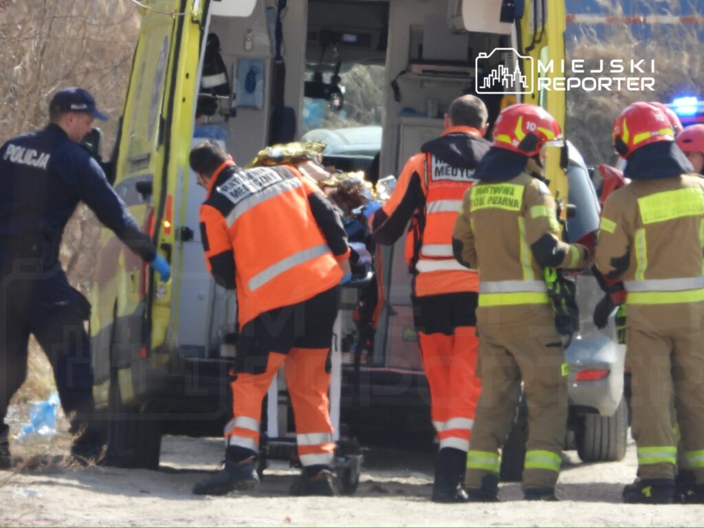 Ratownicy medyczni i strażacy przenoszą poszkodowanego do karetki w terenie zarośniętym trawą.