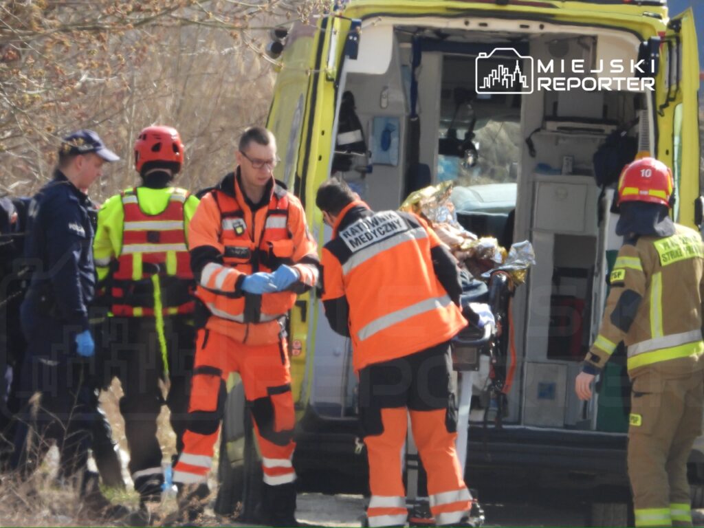 Ratownicy medyczni oraz strażacy udzielają pomocy poszkodowanemu przy ambulansie w terenie.