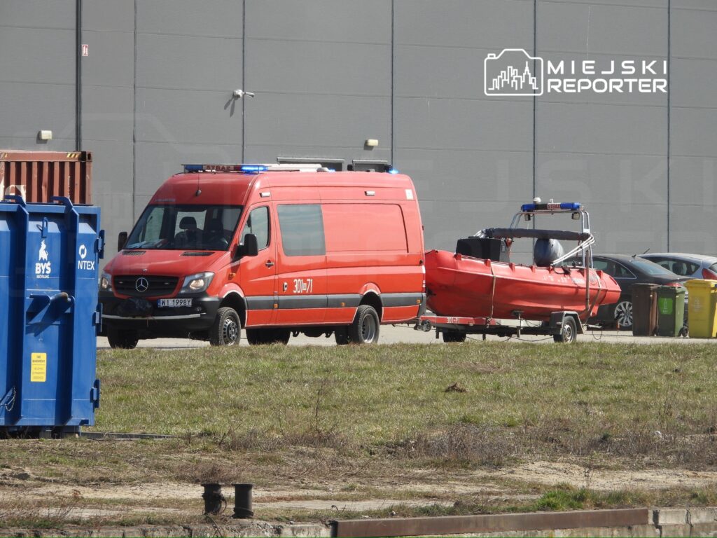 Czerwony samochód ratunkowy z przyczepioną łodzią wędkarską stoi na parkingu obok kontenerów.