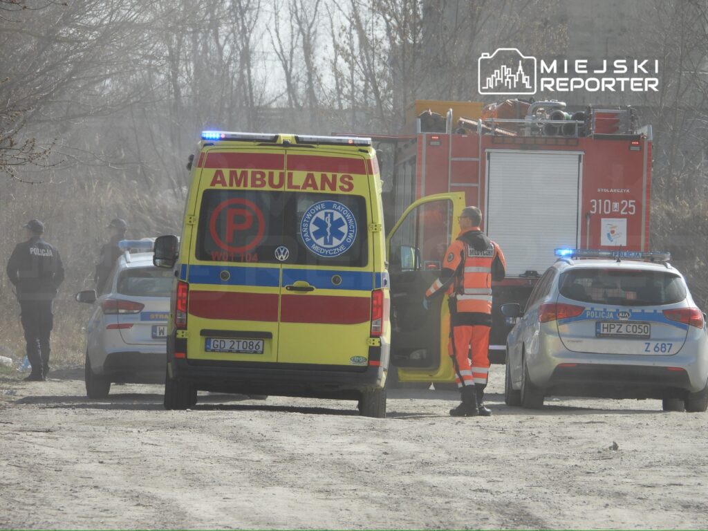 Karetka pogotowia oraz pojazdy policyjne na drodze, w tle straż pożarna i ratownicy medyczni w akcji.