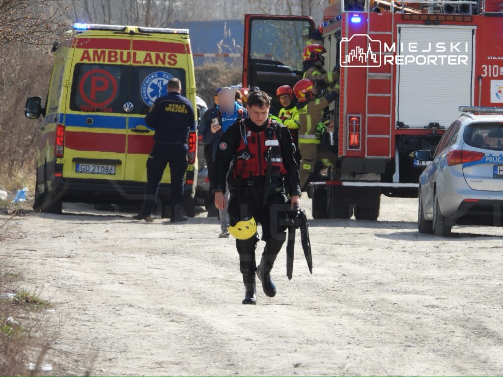 Ratownik w specjalistycznym ubraniu idzie w kierunku pojazdu straży pożarnej, w tle widoczni inni ratownicy.