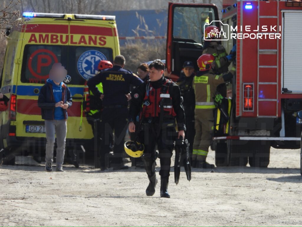 Ratownik w specjalistycznym ubraniu z hełmem, idzie w kierunku karetki, obok grupy strażaków i ratowników medycznych.