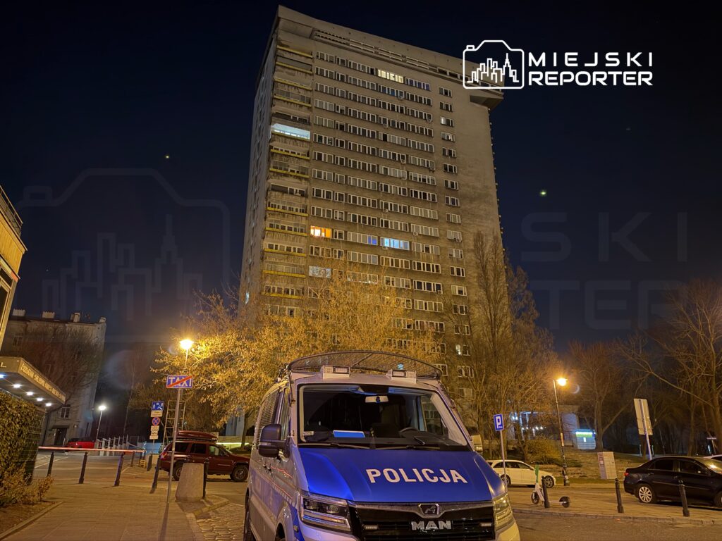 Policyjny radiowóz zaparkowany na ulicy, obok wysokiego bloku mieszkalnego w nocy.