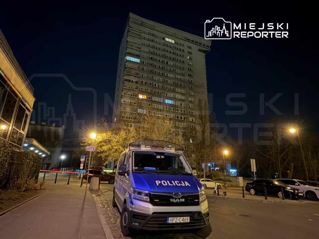 Policyjny radiowóz zaparkowany przy oświetlonym chodniku, obok wysokiego budynku mieszkalnego w nocy.