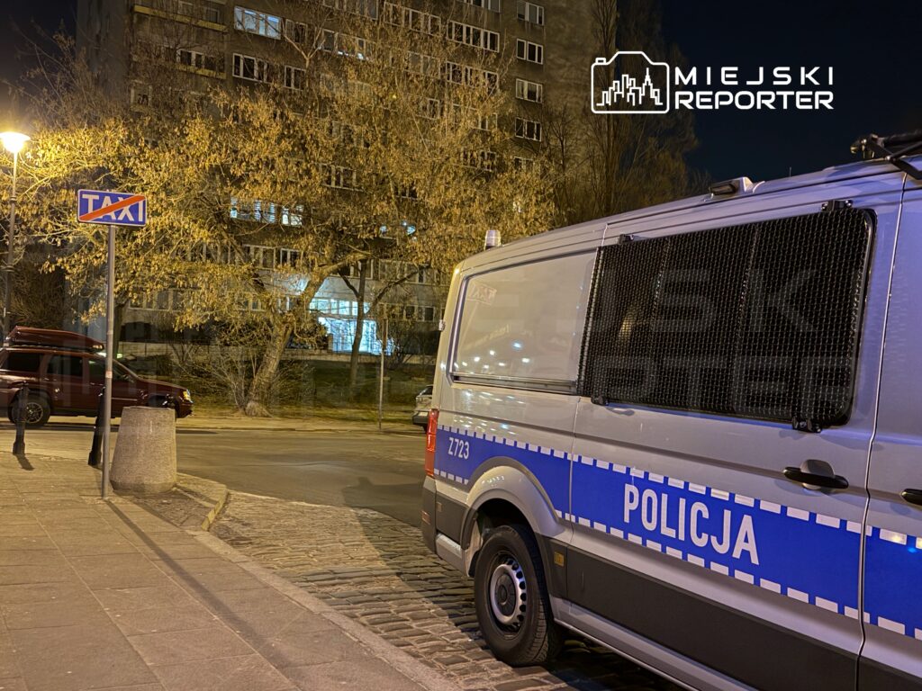 Policyjny samochód zaparkowany przy znaku "TAXI" na oświetlonej ulicy, w tle widać budynek mieszkalny.