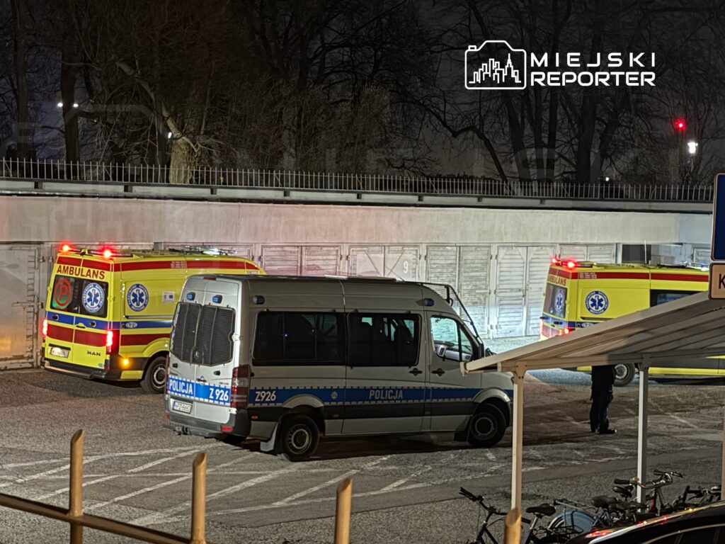 Ambulans i radiowóz policyjny zaparkowane w ciemnym otoczeniu, obok widoczny personel medyczny.