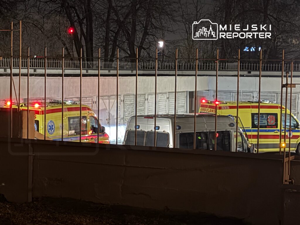 Dwa karetki pogotowia z włączonymi sygnalizatorami świetlnymi stoją na parkingu nocą, otoczone drutem kolczastym.