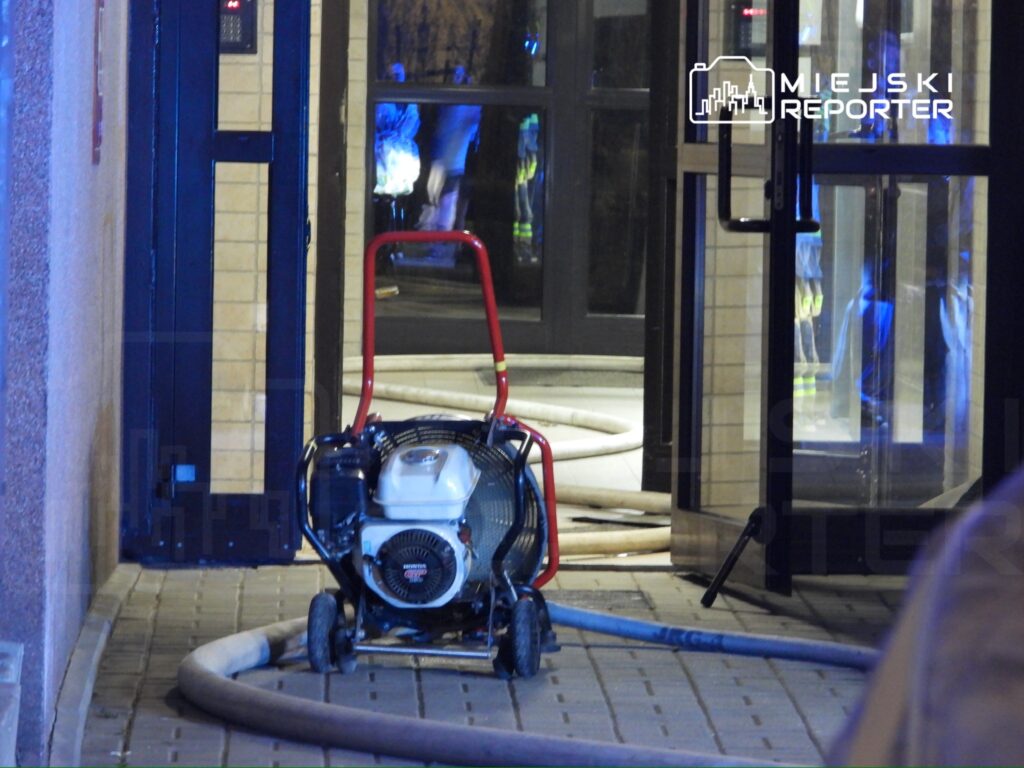 Pompa pożarnicza na wózku stoi na chodniku przed wejściem do budynku, obok węża strażackiego.