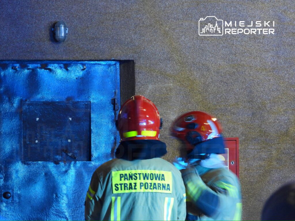 Dwóch strażaków w hełmach omawia sytuację przed niebieską drzwiami w budynku.