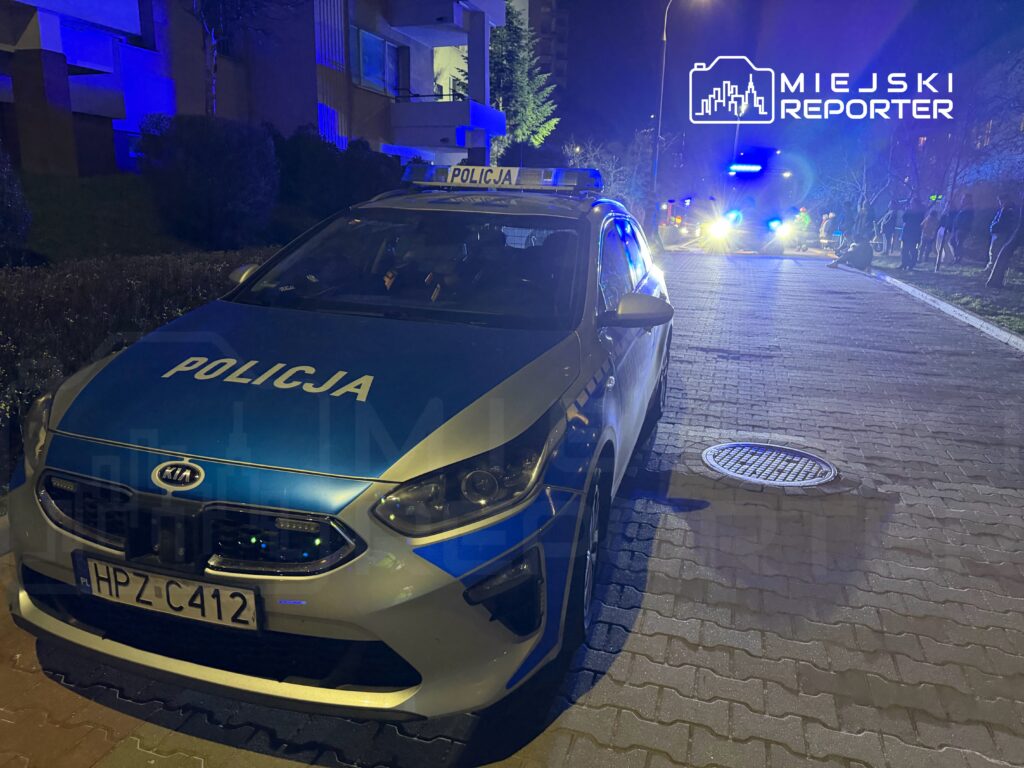 Policyjny samochód z niebieskimi światłami zaparkowany na oświetlonej ulicy, w tle widać grupę ludzi.