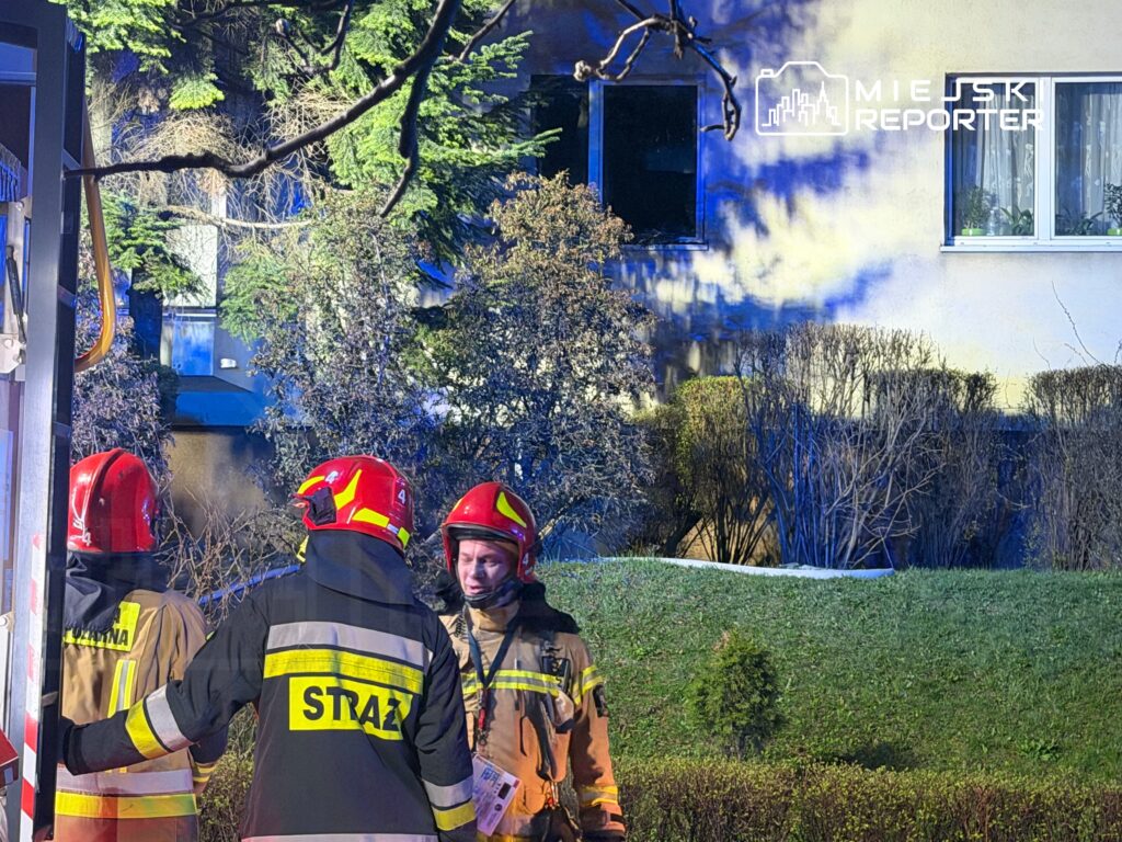 Strażacy w hełmach rozmawiają przed budynkiem, w którym widać dym wydobywający się z okna.