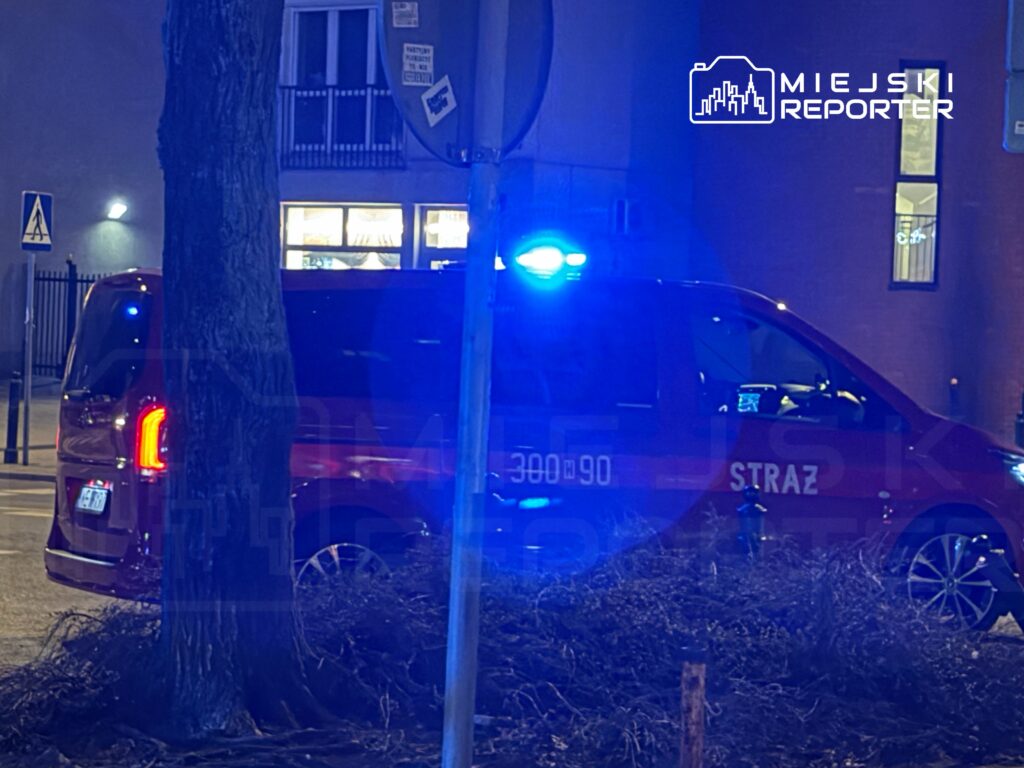 Czerwony samochód strażacki z włączonymi niebieskimi światłami stoi na ulicy obok drzewa.