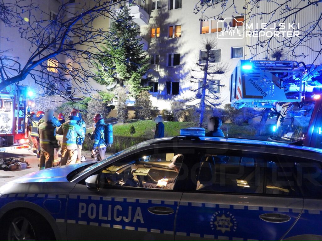 Grupa strażaków w hełmach omawia sytuację przy wozie strażackim, podczas akcji ratunkowej w nocy.
