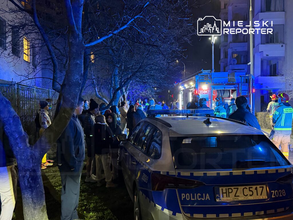 Grupa ludzi obserwująca działania służb ratunkowych przy zaparkowanym radiowozie policyjnym w nocy.