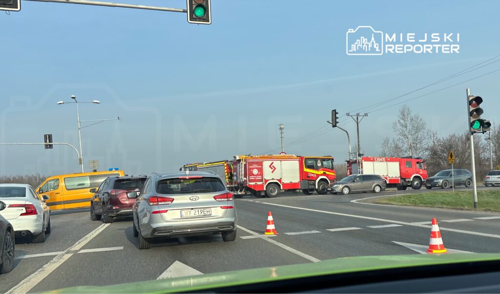 Ruch samochodowy zatrzymany na skrzyżowaniu, obok stoją dwa wozy strażackie z włączonymi sygnałami świetlnymi.