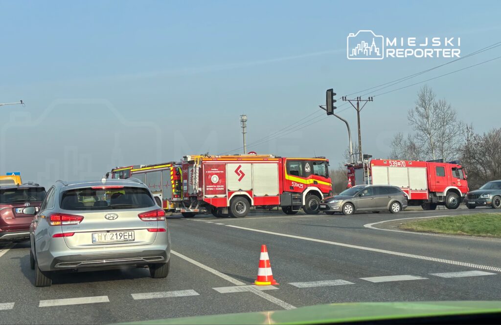 Na skrzyżowaniu zatrzymane pojazdy, w tym dwa wozy strażackie, blokują ruch. W tle widoczny sygnalizator świetlny.