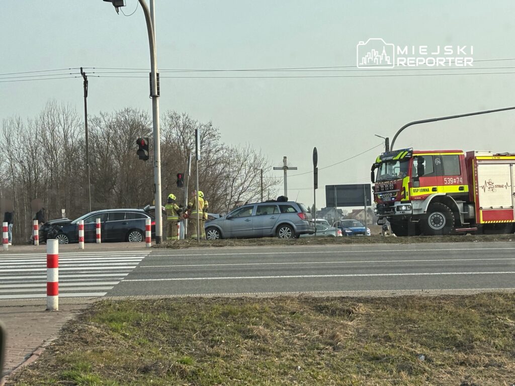 Dwie osoby w uniformach strażackich badają uszkodzone samochody na skrzyżowaniu, obok czeka wóz strażacki.