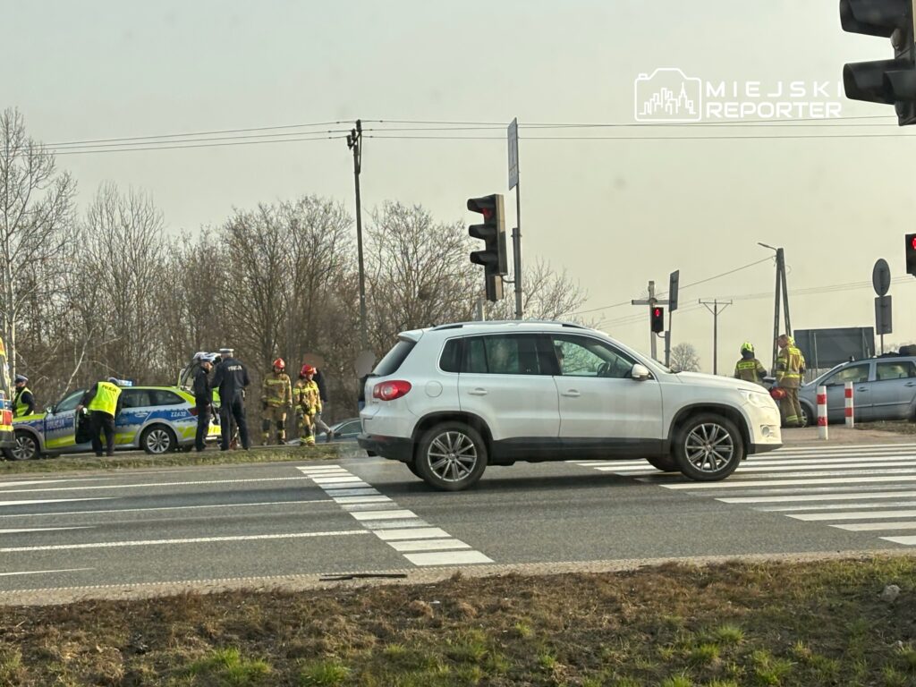 Na skrzyżowaniu policjanci i strażacy udzielają pomocy po wypadku drogowym. W tle widać pojazdy oraz sygnalizację świetlną.