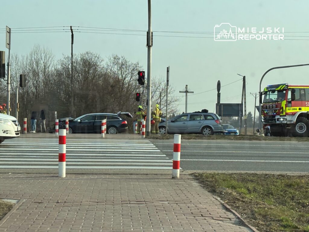 Na skrzyżowaniu stoją dwa samochody, a obok pracują strażacy w żółtych strojach. W tle widoczny jest wóz strażacki.