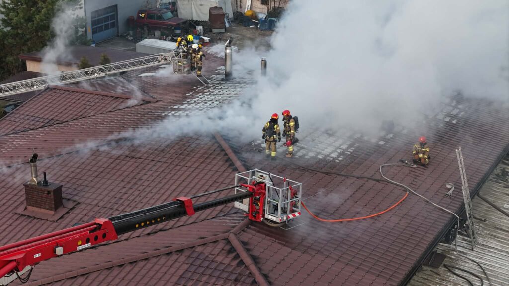 Strażacy w hełmach gaszą pożar na dachu budynku, obok rozprzestrzenia się gęsty dym.