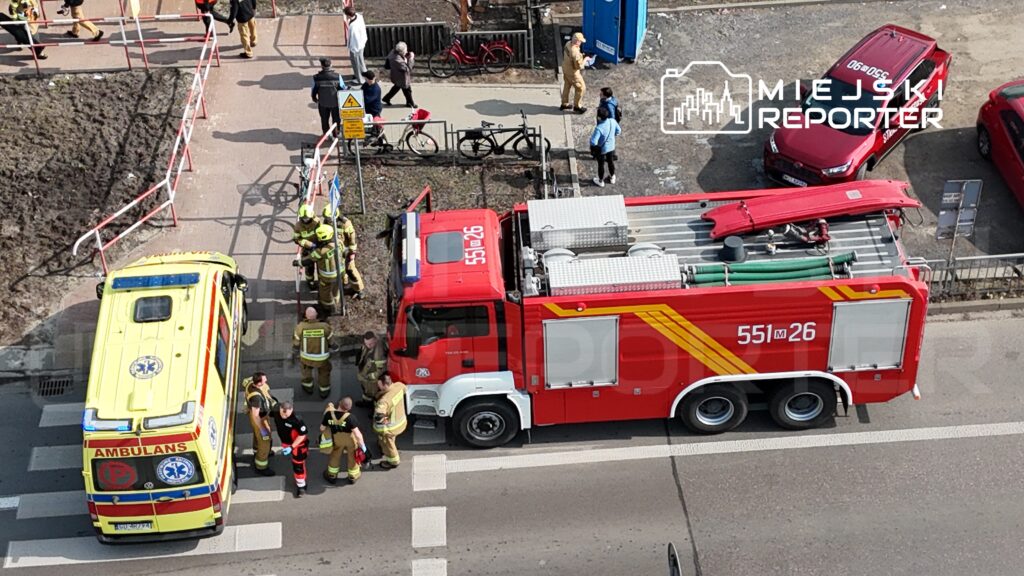 Grupa strażaków w mundurach omawia sytuację obok wozu strażackiego na skrzyżowaniu, obok karetki pogotowia.