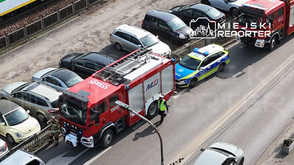 Strażak w mundurze rozmawia z policjantem obok wozu strażackiego i radiowozu, na ulicy otoczonej zaparkowanymi samochodami.