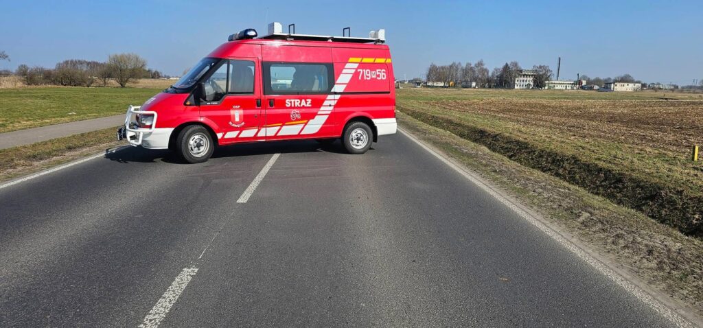 Czerwony wóz strażacki z napisem "STRAŻ" blokujący drogę na wiejskim terenie w słoneczny dzień.