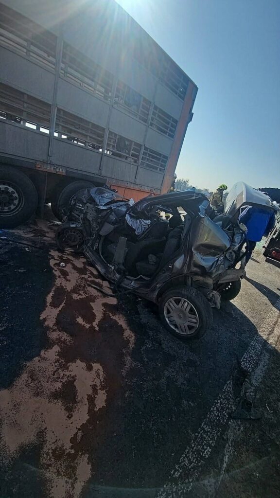Zniszczony samochód osobowy leży na drodze obok ciężarówki, a w tle widać osoby zajmujące się usuwaniem skutków wypadku.
