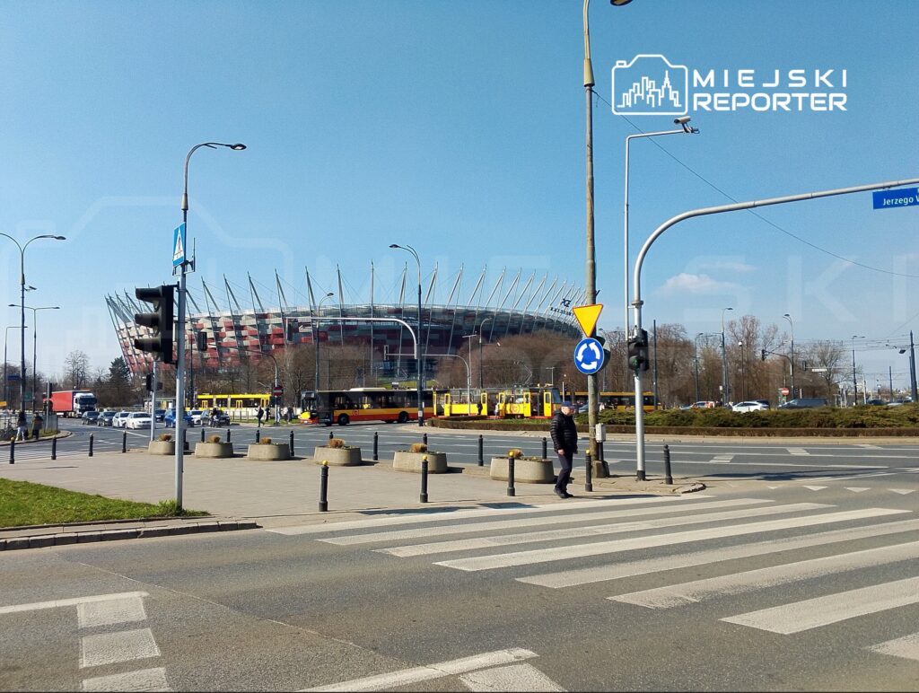 Stadion z charakterystycznymi iglicami w tle, ruchliwa ulica z autobusami i samochodami, pieszy przechodzi przez pasy.