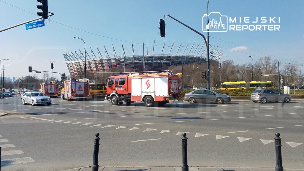 Czerwony wóz strażacki zatrzymany na skrzyżowaniu, w tle stadion z charakterystycznym dachem.