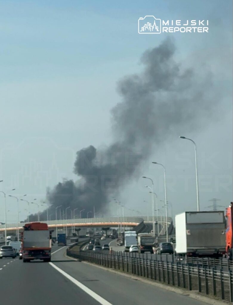 Czarny dym unoszący się nad autostradą, w tle widoczny most i ruchome pojazdy na pasach jezdni.