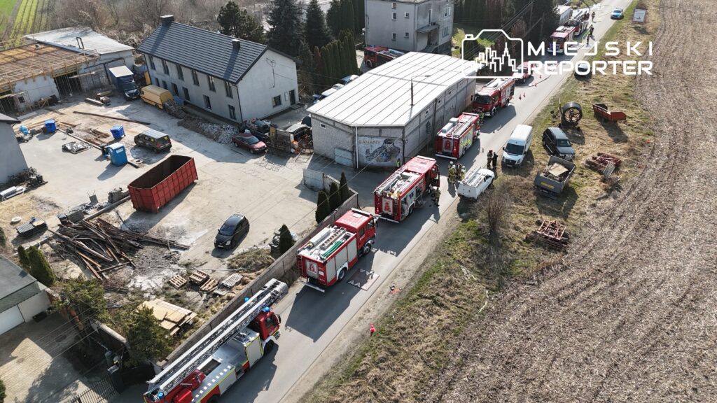 Na drodze stoją samochody strażackie, a obok grupka strażaków rozmawia w pobliżu budynku w trakcie działań ratunkowych.