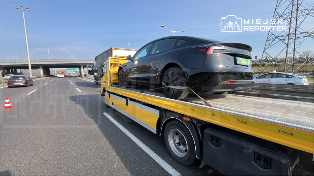 Czarny samochód osobowy na lawecie, zaparkowanej na pasie ruchu, obok innych pojazdów na autostradzie.